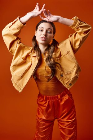Foto de Una mujer con estilo de unos 20 años que lleva una chaqueta amarilla y pantalones naranjas posa sobre un fondo naranja brillante en un entorno de estudio. - Imagen libre de derechos