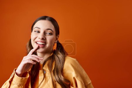 Foto de Una mujer joven y elegante de unos 20 años posa con el dedo en los labios contra un fondo de estudio naranja. - Imagen libre de derechos