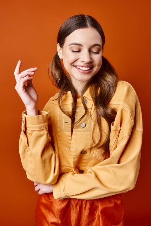 Téléchargez les photos : A stylish young woman in her 20s wearing a yellow shirt and pants poses in a studio with an orange background. - en image libre de droit