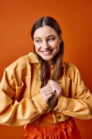 A stylish young woman in her 20s with long hair wearing a vibrant yellow jacket against an orange studio background.