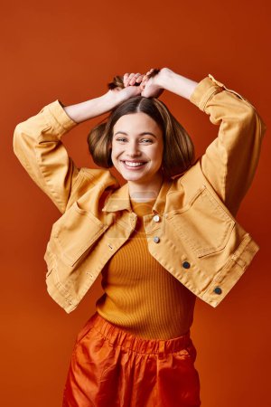 Photo for Young, stylish woman in her 20s, wearing a bright yellow jacket and orange pants, poses in a studio setting with an orange background. - Royalty Free Image