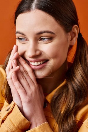 Foto de Una mujer joven y elegante de unos 20 años, sonriendo brillantemente con las manos en la cara contra un fondo de estudio naranja. - Imagen libre de derechos