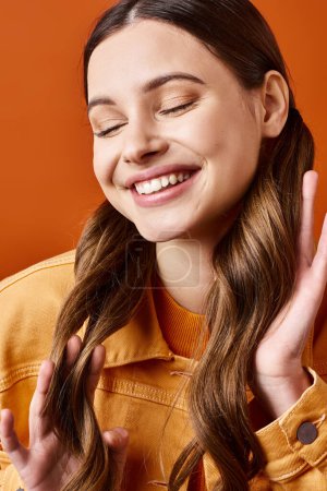 Foto de Una joven y elegante mujer de unos 20 años sonríe brillantemente mientras mantiene sus manos juntas en una expresión alegre, sobre un vibrante fondo naranja. - Imagen libre de derechos
