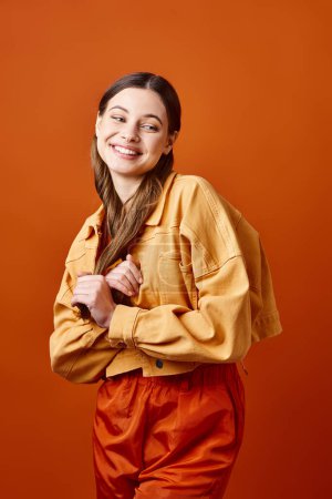 Foto de Una joven con estilo de unos 20 años, de pie con confianza con los brazos cruzados y una sonrisa brillante, contra un fondo de estudio naranja. - Imagen libre de derechos