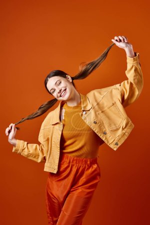 Eine stylische junge Frau in ihren Zwanzigern in gelber Jacke und Hose posiert vor orangefarbenem Hintergrund in einem Studio-Setting.