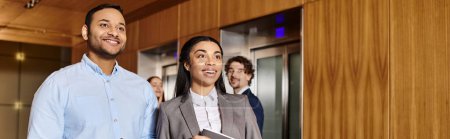 A man and a woman, an interracial group of business professionals, stand in front of an elevator.
