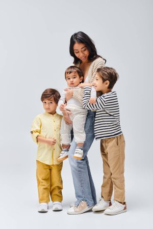 Una joven madre asiática sosteniendo tiernamente a sus hijos, parada cerca en un estudio sobre un fondo gris.