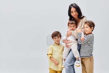 A young Asian mother tenderly cradles children in her arms, conveying love and protection in a studio setting on a grey background.