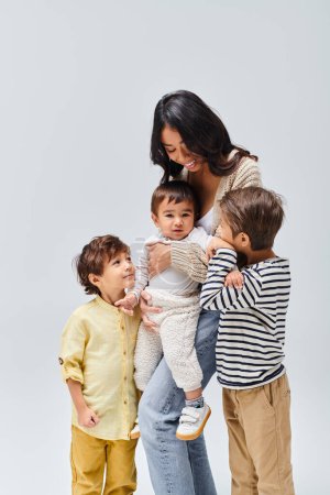 Asian mother tenderly holds her baby while two small children look on, creating a heartwarming family moment in a studio.