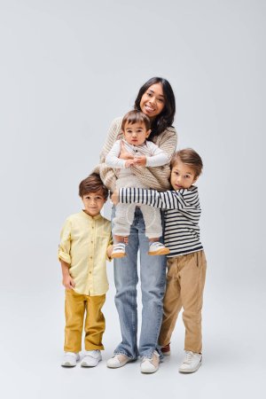 A young Asian mother stands on a grey background, holding her small children in her arms.