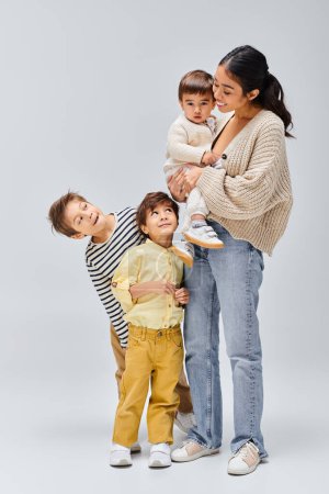 A young Asian mother stands next to her children, in a studio setting with a grey background.