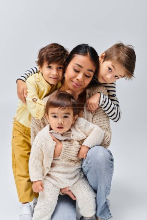 Eine junge asiatische Mutter und ihre drei Kinder posieren freudig für ein Porträt in einem Studio vor grauem Hintergrund.