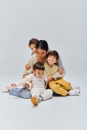 Photo for A young Asian mother sits on the ground with her children in a studio setting on a grey background. - Royalty Free Image