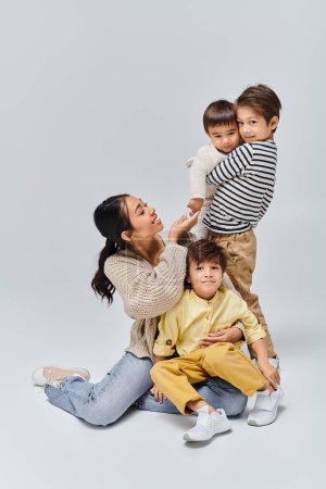 A young Asian mother sits on the ground, warmly embracing her children in a studio setting with a grey background.