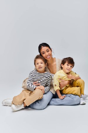 A young Asian mother sits peacefully on the ground with her two children in a studio setting on a grey background.