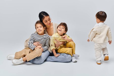 Photo for A young Asian mother sits on the ground with her children in a studio setting against a grey background, bonding closely. - Royalty Free Image