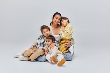 Eine junge asiatische Mutter sitzt mit Kindern auf dem Boden in einem Studio vor grauem Hintergrund.