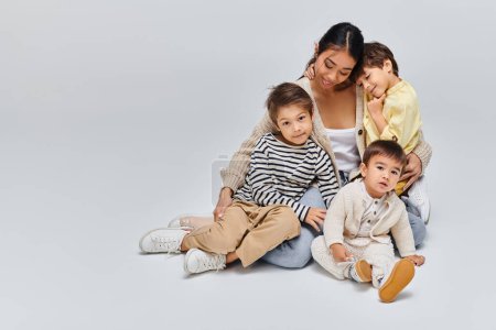 Foto de Una joven madre asiática se sienta en el suelo con sus hijos en un sereno estudio sobre un fondo gris. - Imagen libre de derechos
