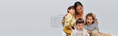 Photo for A young Asian mother sitting on a chair, lovingly embracing her children in a studio setting against a grey background. - Royalty Free Image