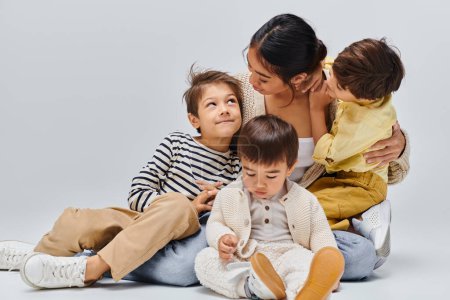 Foto de Una madre asiática se sienta en el suelo con sus hijos, compartiendo un abrazo amoroso en un estudio sobre un fondo gris. - Imagen libre de derechos