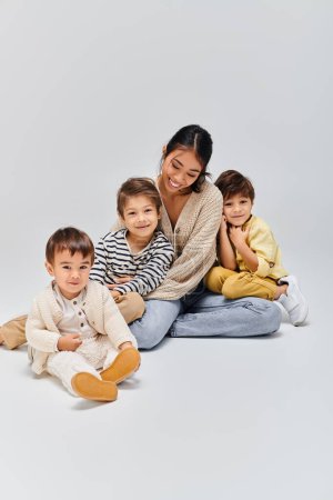 Foto de Una joven madre asiática se sienta en el suelo con sus hijos en un estudio sobre un fondo gris. - Imagen libre de derechos