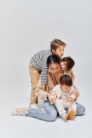 Eine junge asiatische Mutter und ihre Kinder sitzen übereinander in einem Studio vor grauem Hintergrund.
