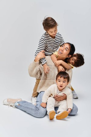 Une jeune mère asiatique et ses enfants s'assoient les uns sur les autres, créant une pyramide humaine dans un studio sur fond gris.
