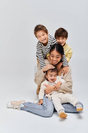 Téléchargez les photos : A young Asian mother sits on the floor with her children in a studio setting against a grey background. - en image libre de droit