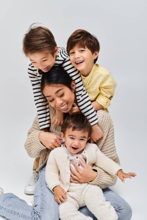 Eine junge asiatische Mutter und ihre Kinder erschaffen eine einzigartige Menschenpyramide, die in einem Atelier vor grauem Hintergrund übereinander sitzt.