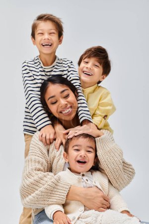 Foto de Una joven madre asiática y sus hijos se apilan juguetonamente uno encima del otro en un entorno de estudio con un fondo gris. - Imagen libre de derechos