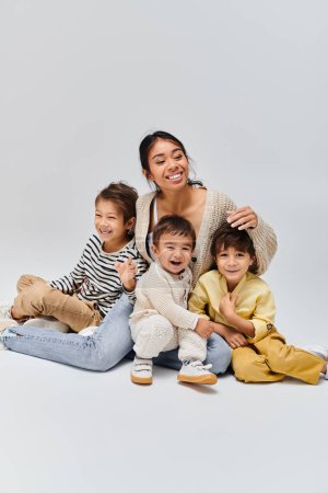 Photo for A young Asian mother sits peacefully on the ground with her kids in a studio setting against a grey background. - Royalty Free Image