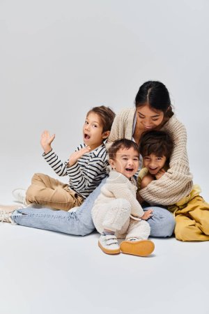 Téléchargez les photos : Une jeune mère asiatique assise par terre entourée de ses enfants dans un studio sur fond gris. - en image libre de droit