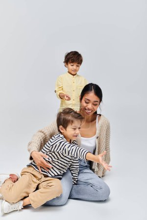 Téléchargez les photos : A young Asian mother sits on the ground with children beside her in a studio setting against a grey background. - en image libre de droit