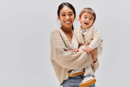 Foto de Una joven madre asiática sostiene tiernamente a su bebé en brazos en un estudio sobre un fondo gris. - Imagen libre de derechos