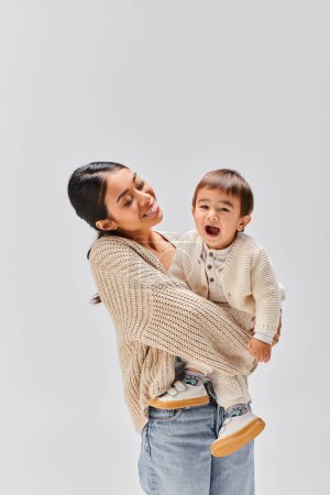 Une jeune mère asiatique tient tendrement son bébé dans ses bras, montrant amour et soins dans un cadre de studio sur un fond gris.