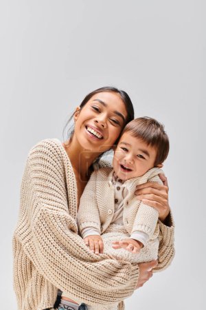 Una joven madre asiática sostiene tiernamente a su hijo en brazos contra un suave fondo gris del estudio.