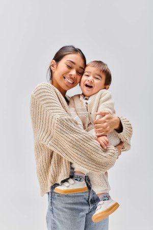 Téléchargez les photos : Une jeune mère asiatique berçant tendrement son bébé dans ses bras en studio sur fond gris. - en image libre de droit