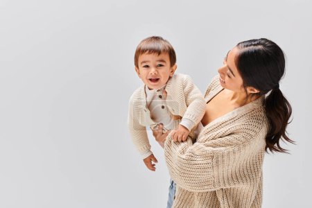 Une jeune mère asiatique tient tendrement son bébé dans ses bras dans un décor de studio sur fond gris.