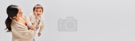 Photo for A young Asian mother lovingly holds her small child in her arms in a studio setting against a grey background. - Royalty Free Image