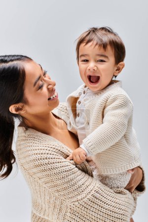 A young Asian mother tenderly holds her baby in her arms in a studio setting against a grey background.