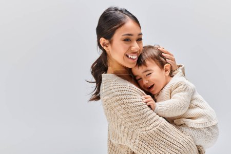 Photo for A young Asian mother tenderly embracing her child in a studio setting against a grey background. - Royalty Free Image