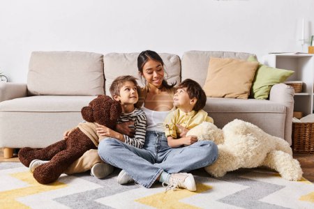 Eine junge asiatische Mutter sitzt mit ihren beiden Kindern und einem Teddybär im heimischen Wohnzimmer auf dem Fußboden.