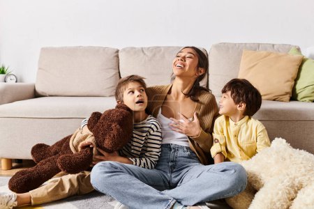 A young Asian mother sitting on the floor with her two children and a teddy bear, creating a cozy family moment.