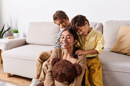 Foto de A young Asian mother sits on a couch, holding a teddy bear in her arms, surrounded by her little sons in a cozy living room. - Imagen libre de derechos