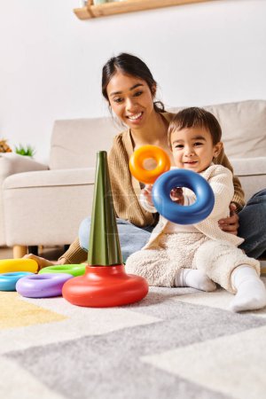 Eine junge asiatische Mutter und ihr kleiner Sohn spielen fröhlich mit Spielzeug auf dem Fußboden in ihrem gemütlichen Wohnzimmer.