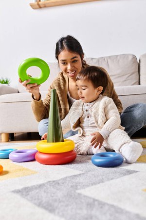 Téléchargez les photos : Une jeune mère asiatique interagit joyeusement avec son petit fils sur le sol de leur salon à la maison. - en image libre de droit