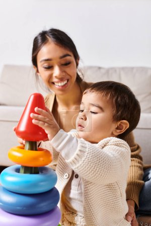 A young Asian mother joyfully plays with her little son on the floor of their cozy living room.