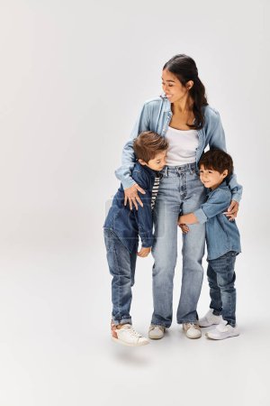 Eine junge asiatische Mutter und ihre kleinen Söhne, alle in Jeanskleidung, posieren in einem grauen Studio für ein Foto.