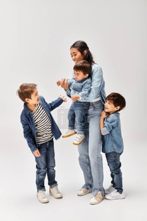 Foto de Una joven madre asiática y sus hijos pequeños, vestidos de mezclilla, pasando un rato alegre y juguetón juntos en un estudio gris. - Imagen libre de derechos