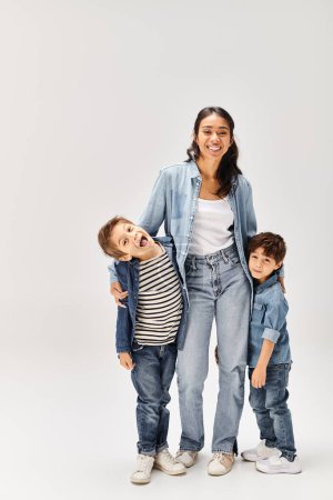 Eine junge asiatische Mutter und ihre kleinen Söhne, alle in Jeans-Outfits, posieren gemeinsam für ein Porträt in einem grauen Studio.